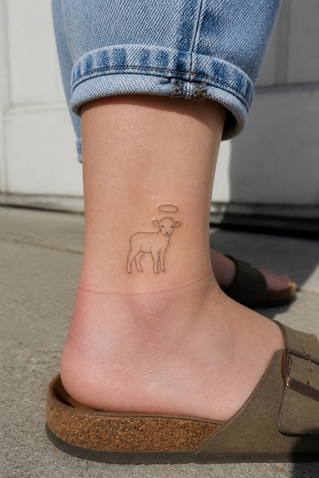 Outline tattoo of a lamb with a halo on the ankle in fine line style.