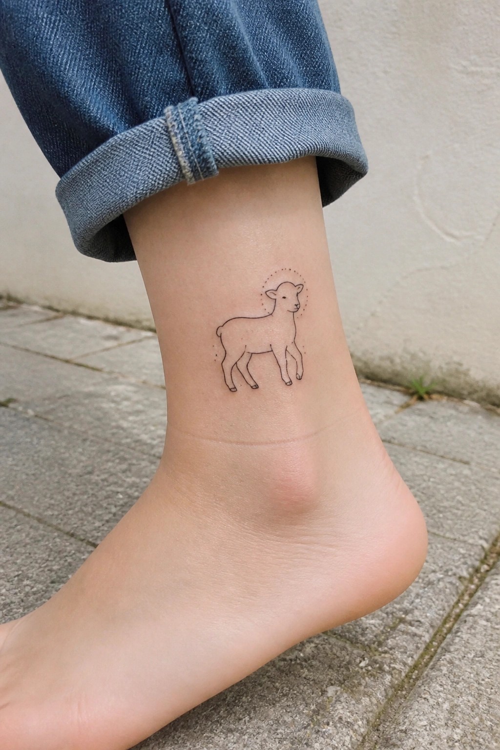 Bare ankle with simple lamb tattoo and dotted halo, cuffed blue jeans on pavement