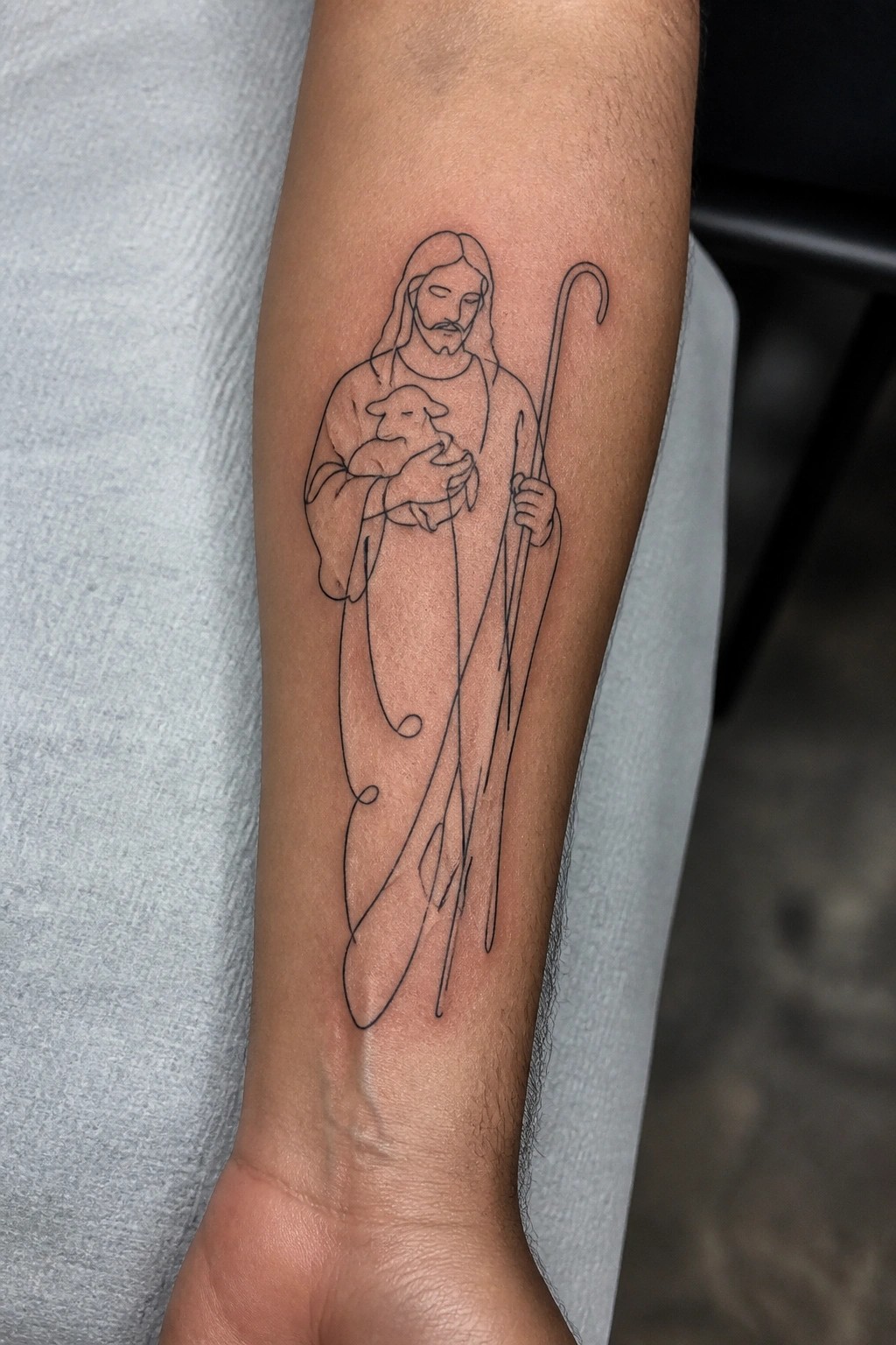 Minimalist line art tattoo of Jesus holding a lamb and shepherd's staff on the inner forearm