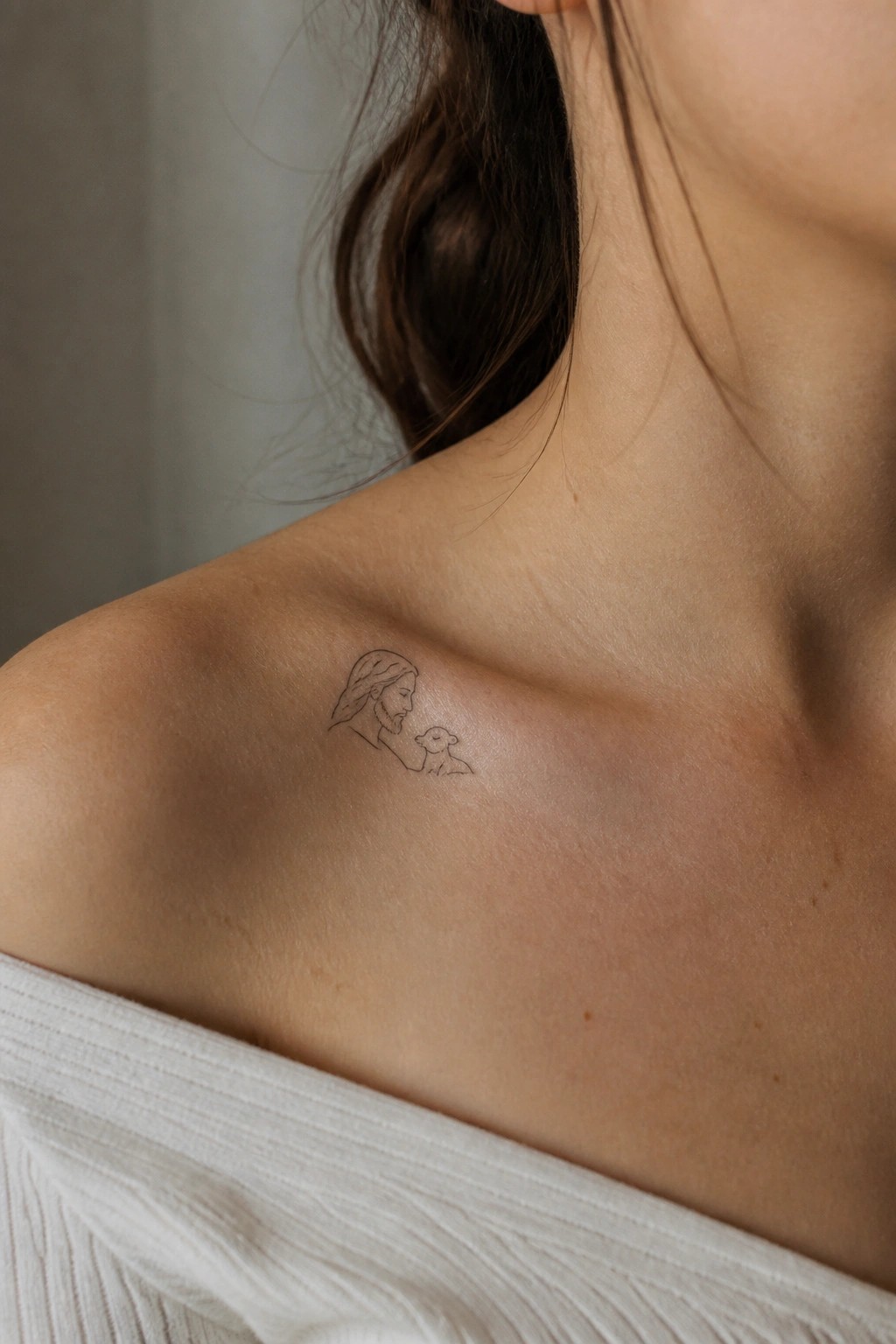 Small line-art tattoo of Jesus and lamb on woman's bare shoulder with white top