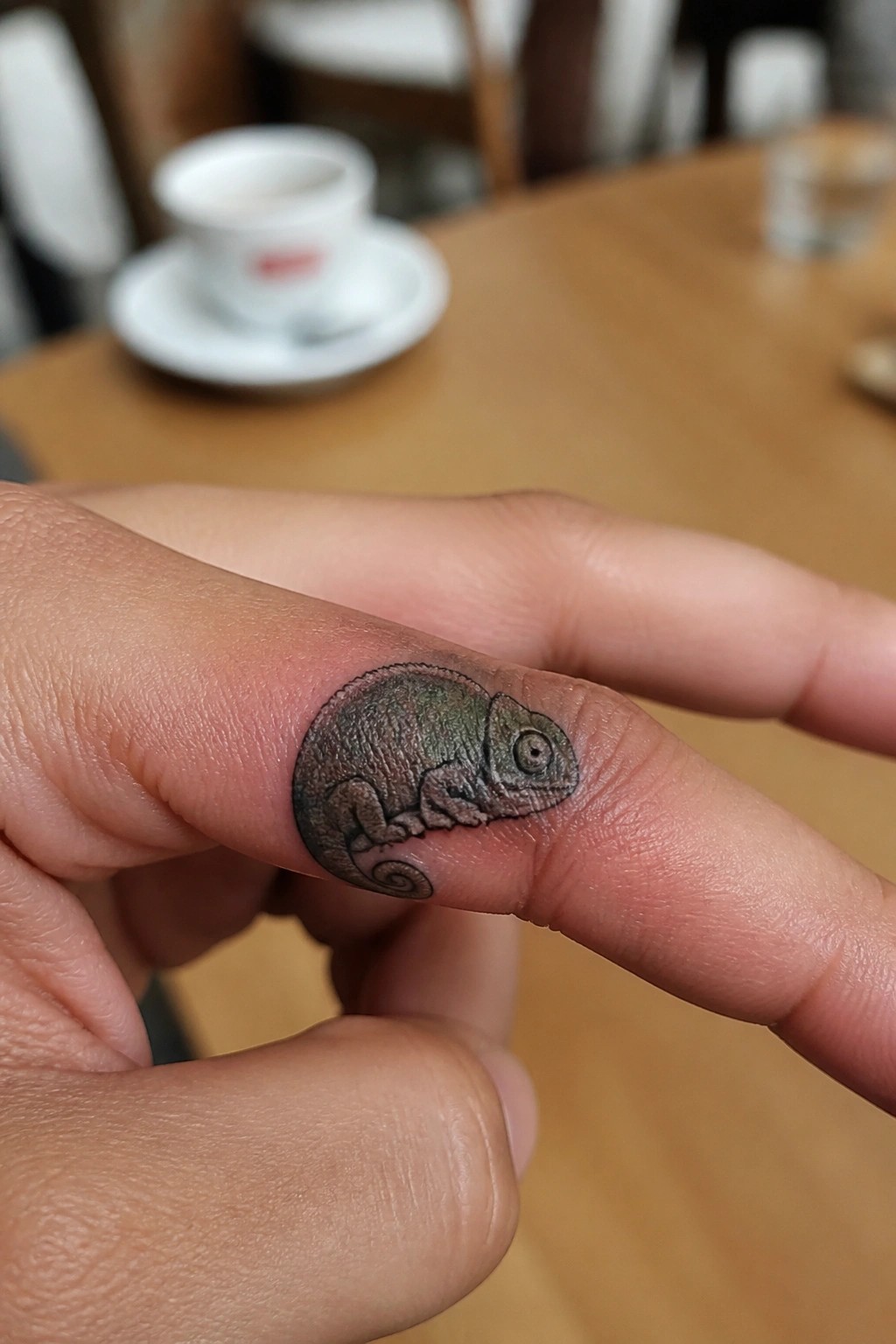 Small realistic chameleon tattoo curled on a finger with fine shading and detail