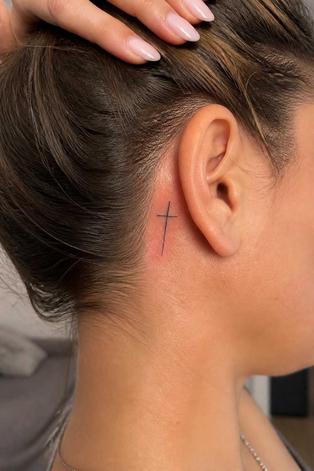 Minimalist black line cross tattoo placed directly behind a woman's ear on the side of the head.