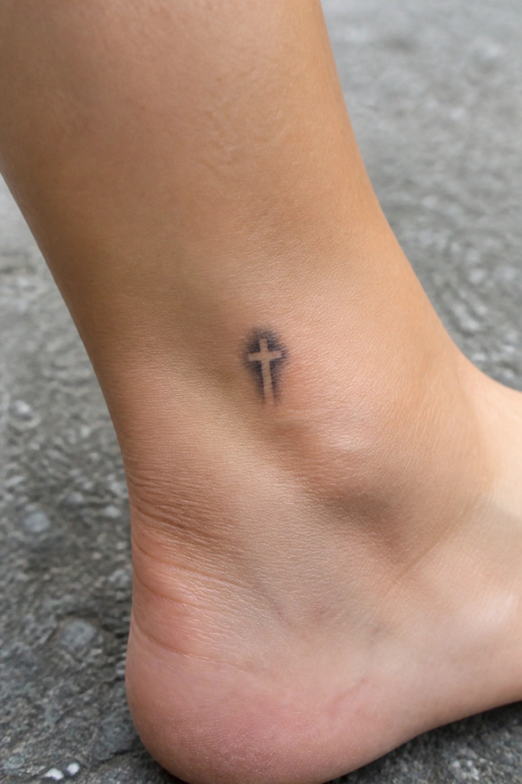 Small black cross tattoo on inner ankle above bare foot on pavement