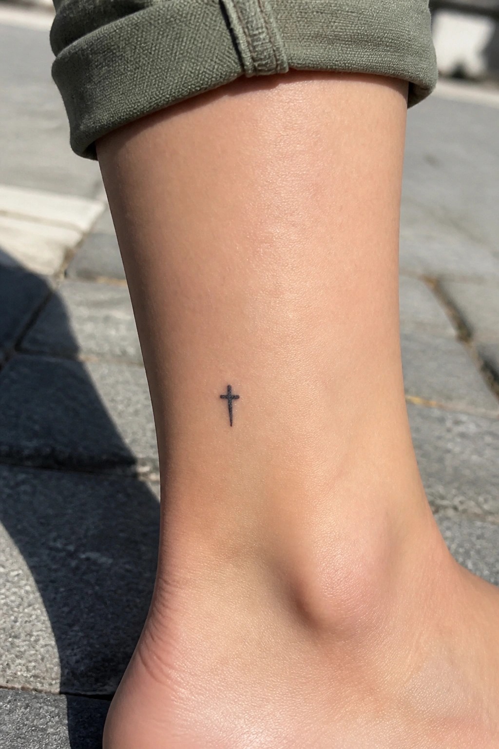 Small black cross tattoo on the inner ankle of a person wearing rolled green pants.