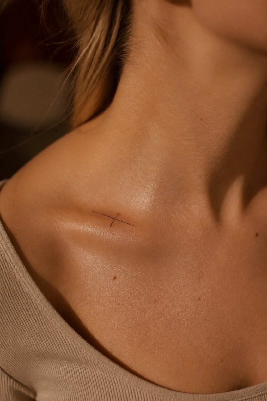 Small cross tattoo on fair skin near collarbone, beige top visible in warm light