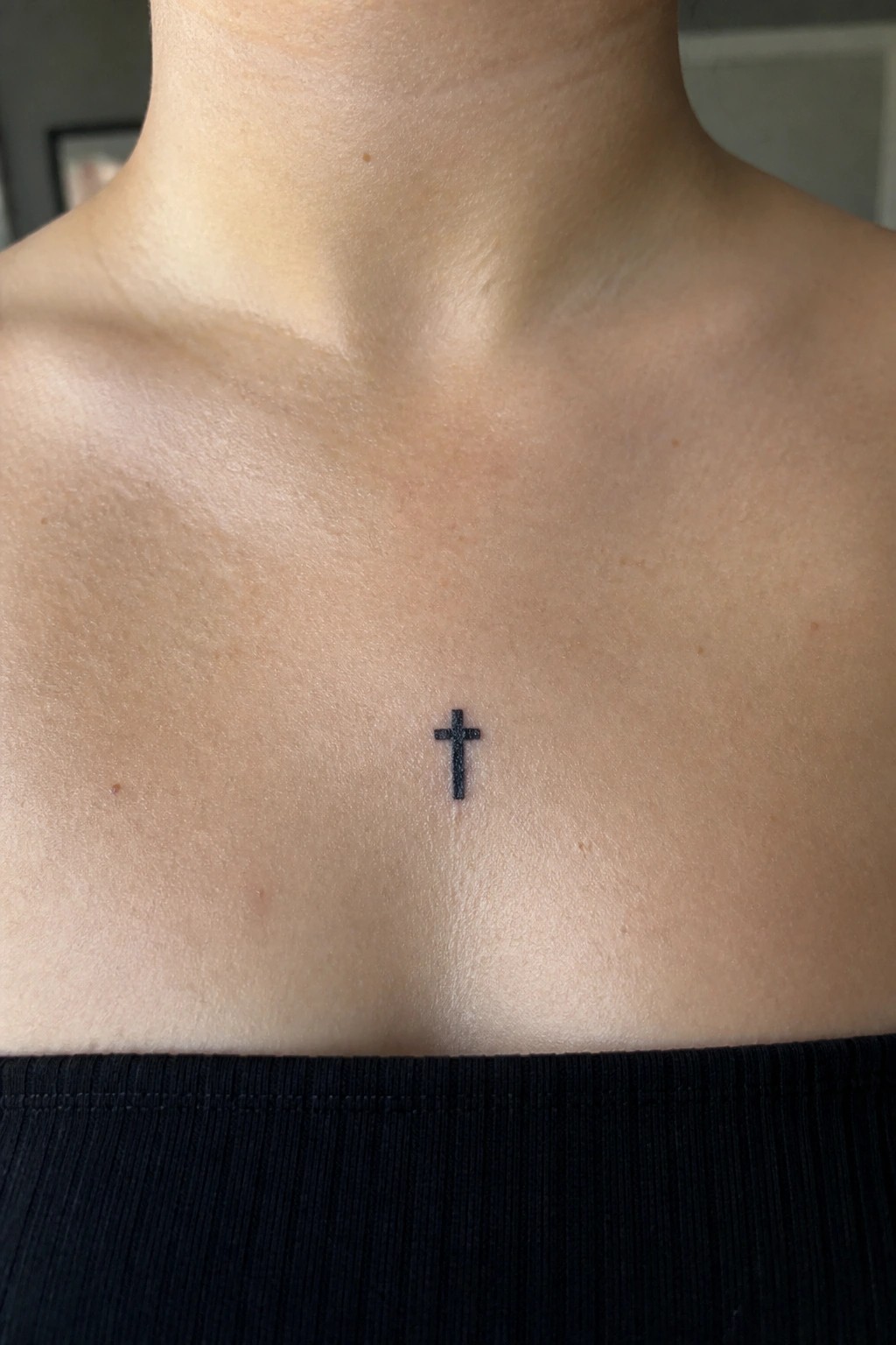 Small black cross tattoo centered on light-skinned upper chest above black top