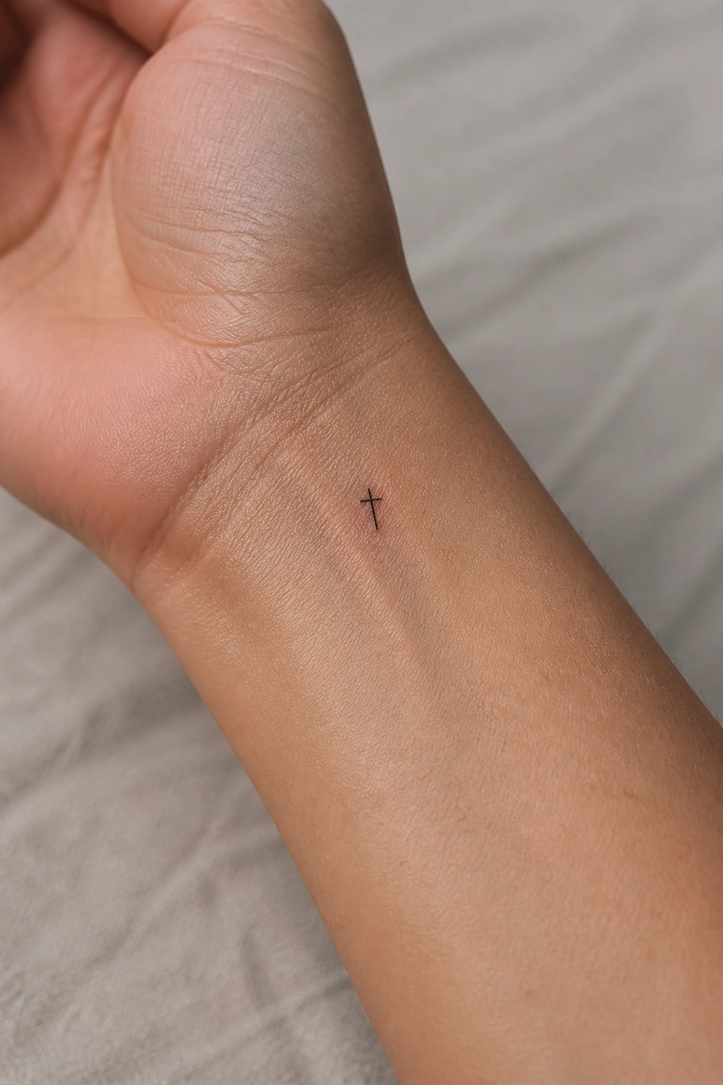 Small fine line cross tattoo on the inner wrist of a hand resting on fabric