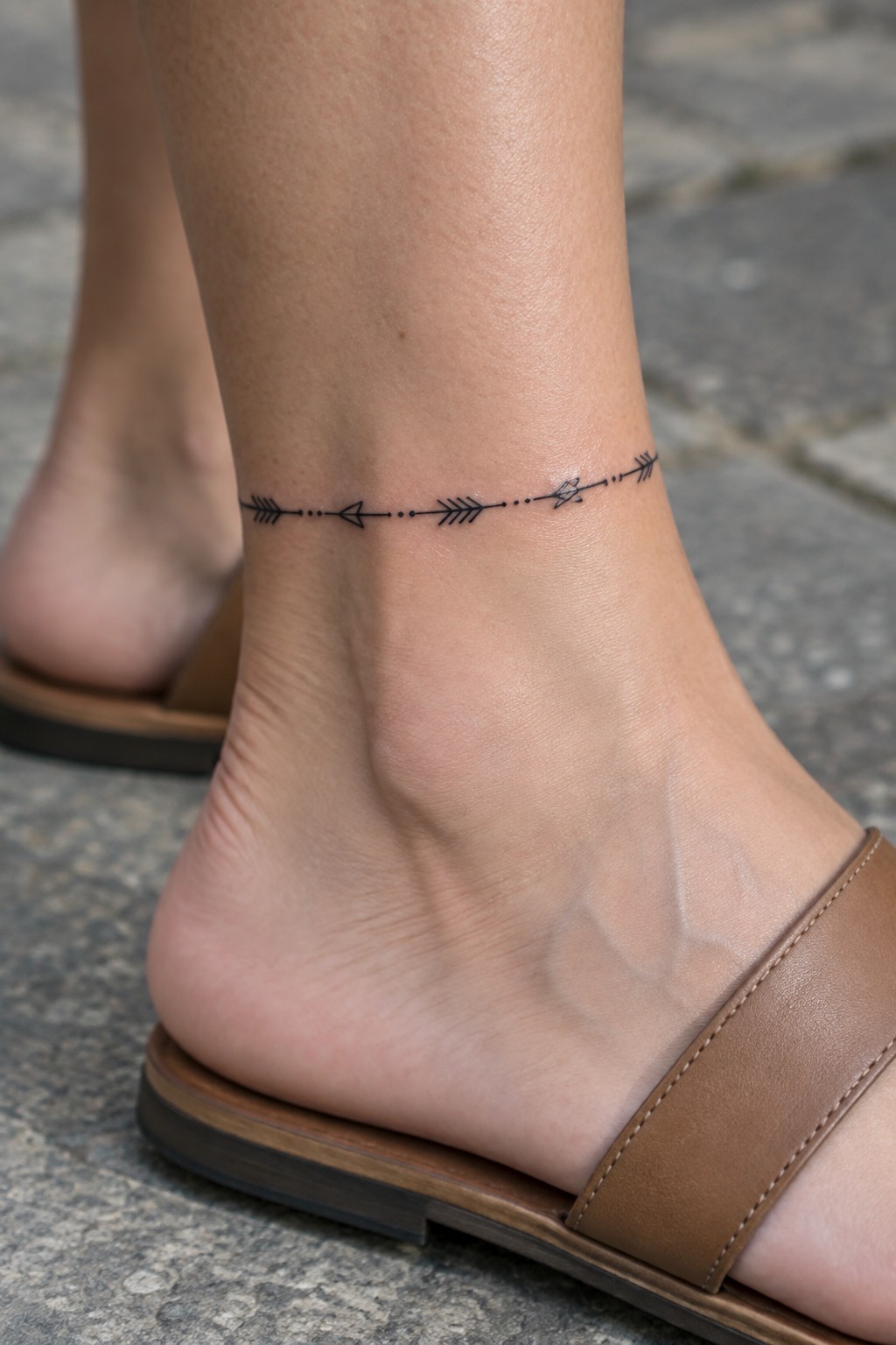 Ankle with black arrow tattoo above brown leather sandal on pavement.