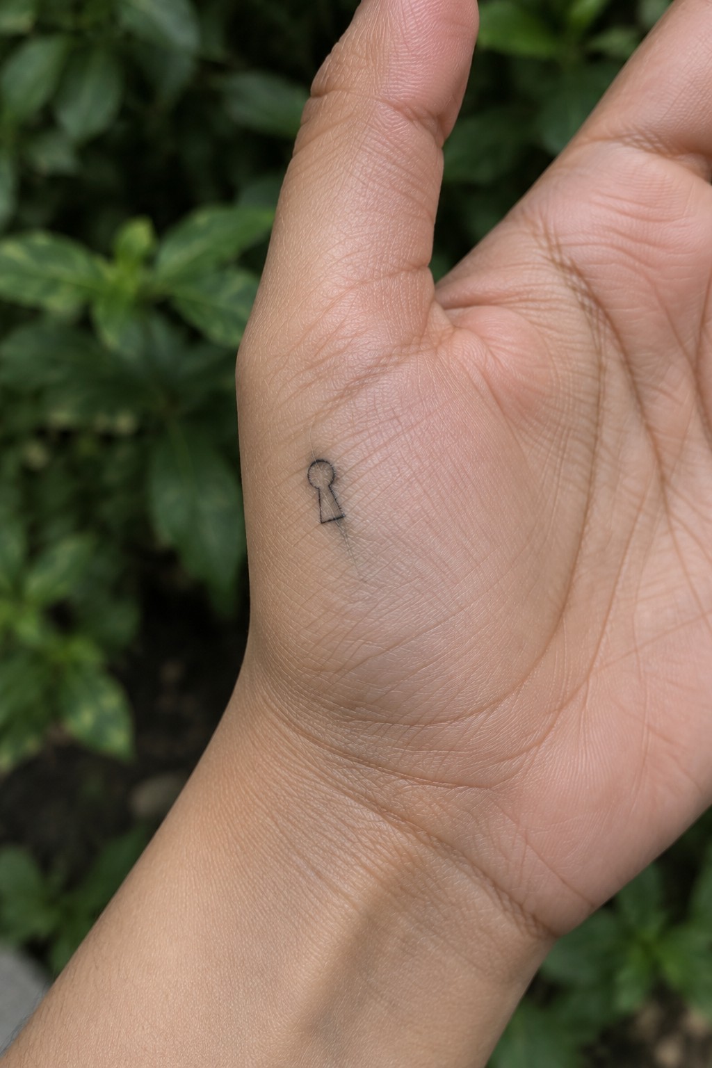 Close-up of light-skinned hand with small black keyhole tattoo on palm, green foliage background.