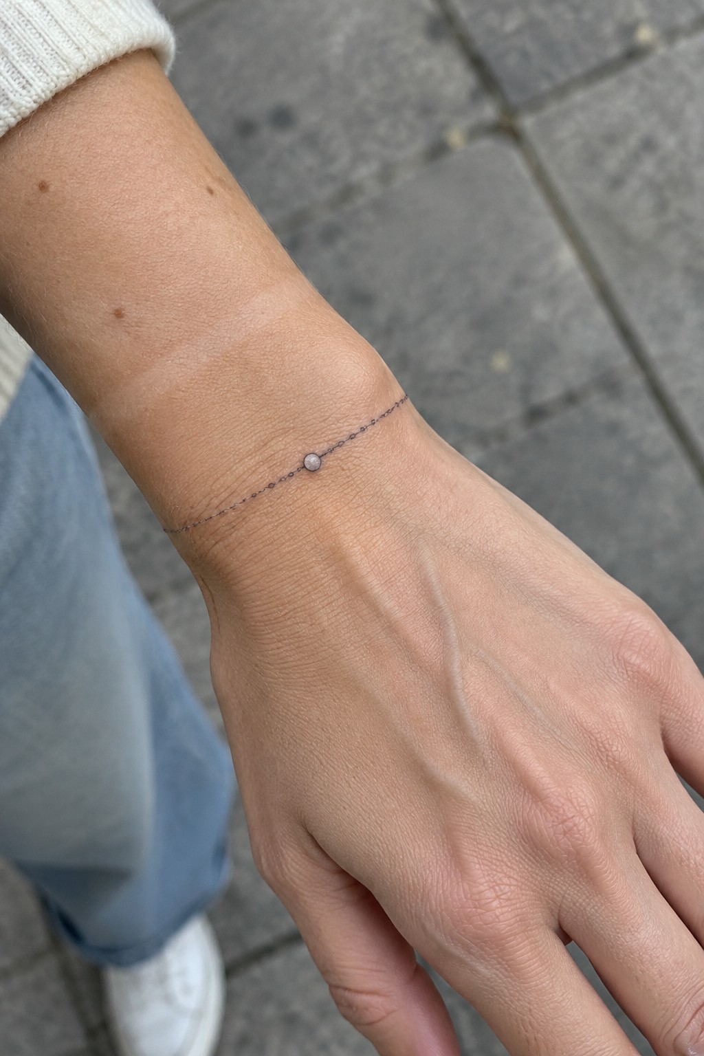 Fine line black ink chain bracelet tattoo with a single bead centered on a woman's wrist.