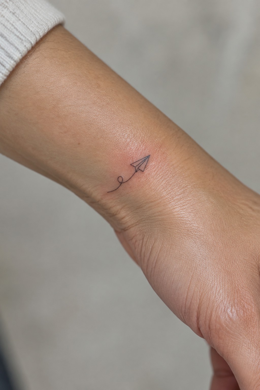 Paper airplane tattoo with trail on wrist beside white sweater cuff