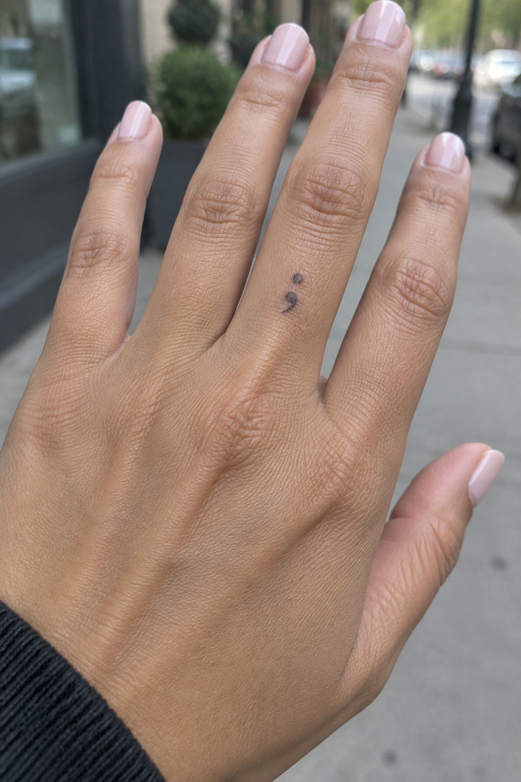Hand with light pink nails and black sleeve, showing semicolon tattoo on ring finger.