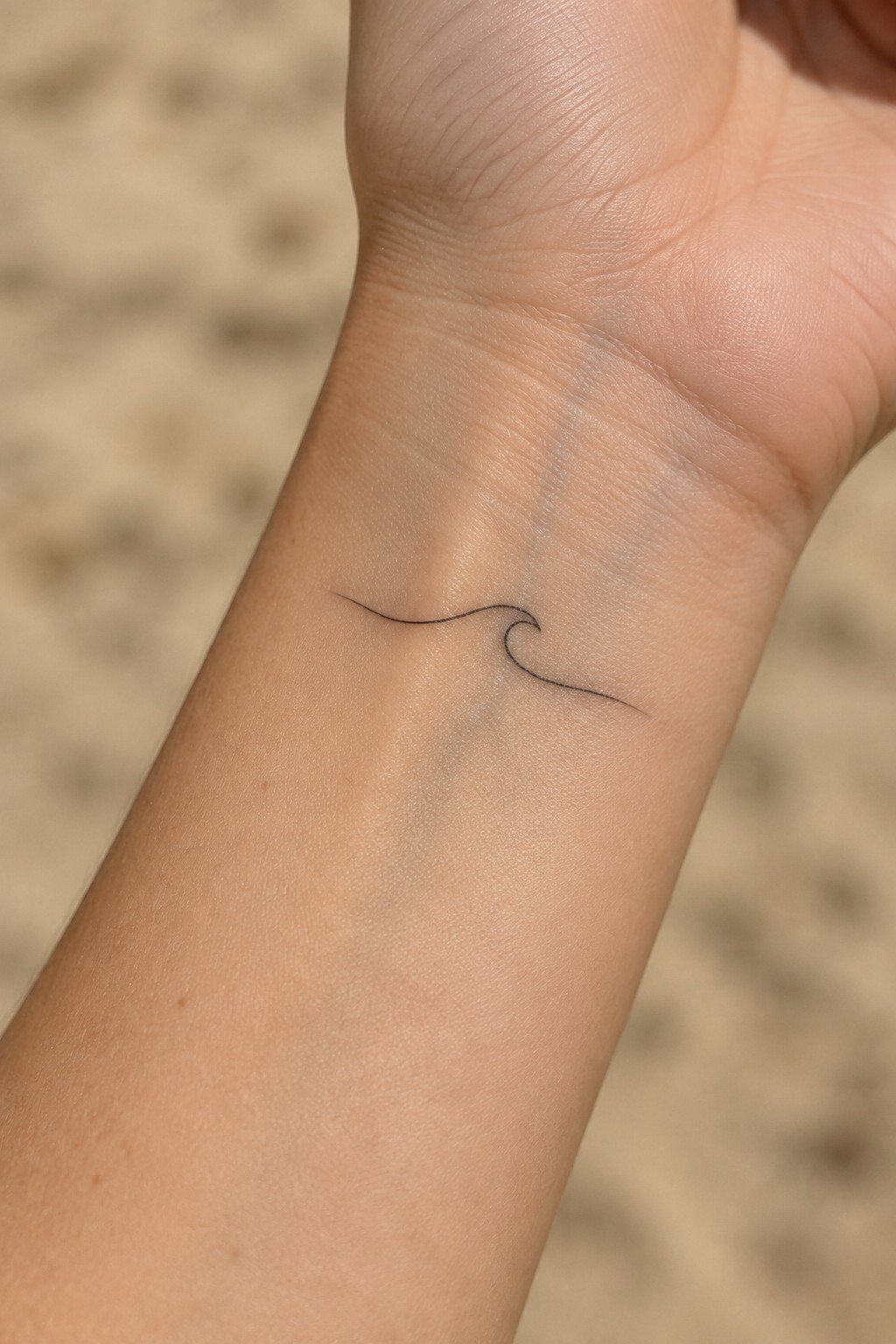 Small black wave tattoo on light-skinned wrist over blurred beach sand