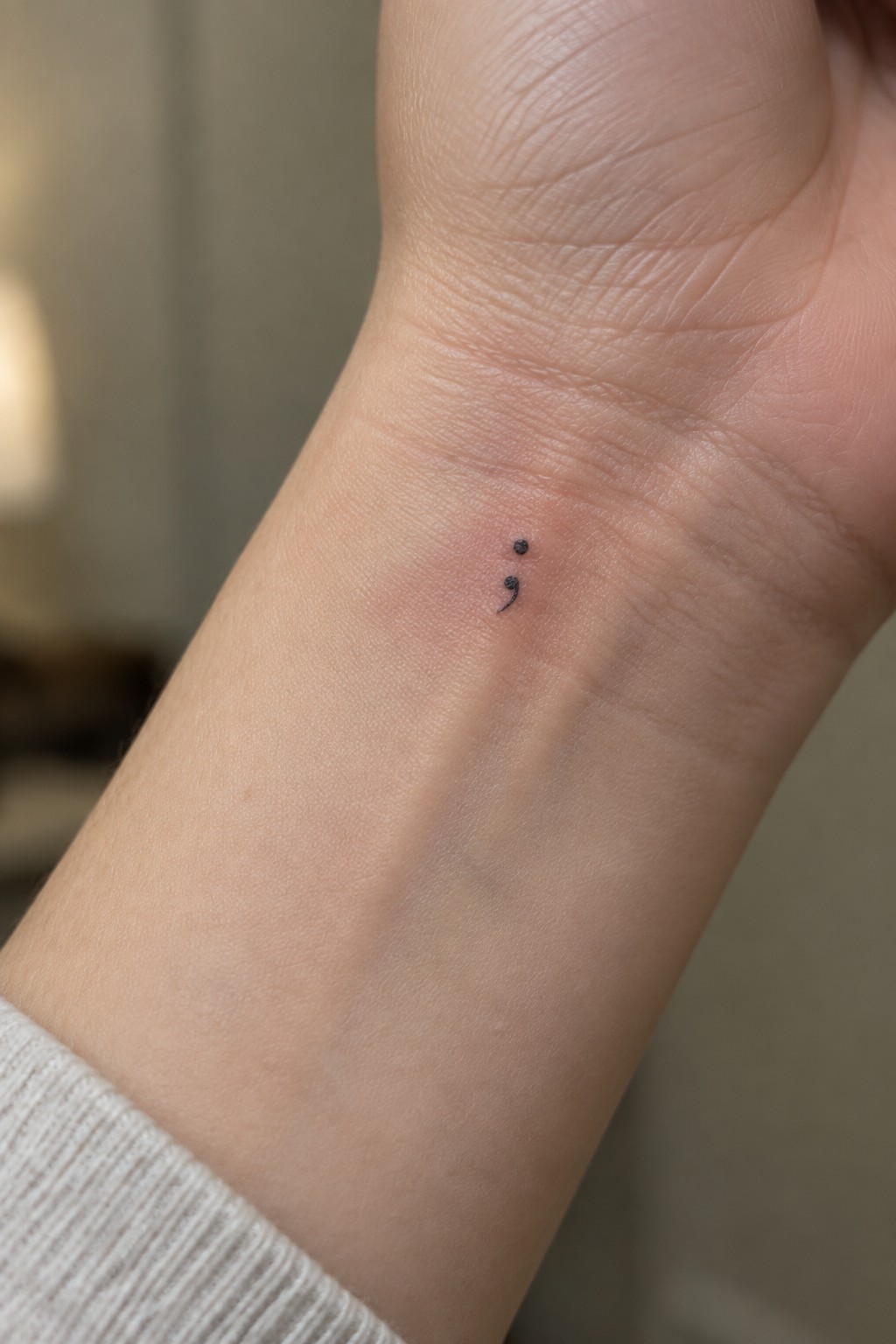 Light-skinned wrist with small black semicolon tattoo and white sweater cuff