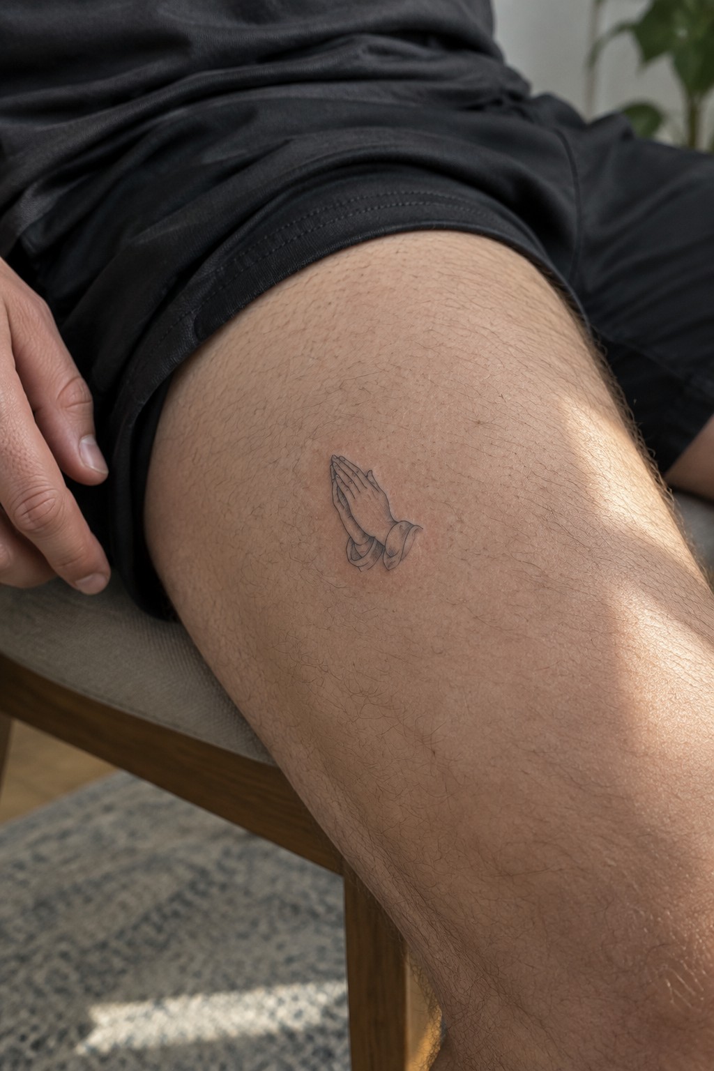 Small black line drawing of praying hands tattooed on a man's upper thigh