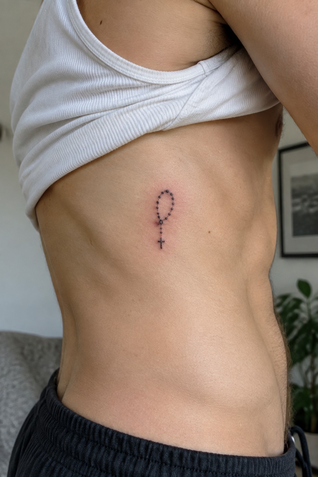 Small black rosary tattoo with beads and cross placed on the side of a man's torso.