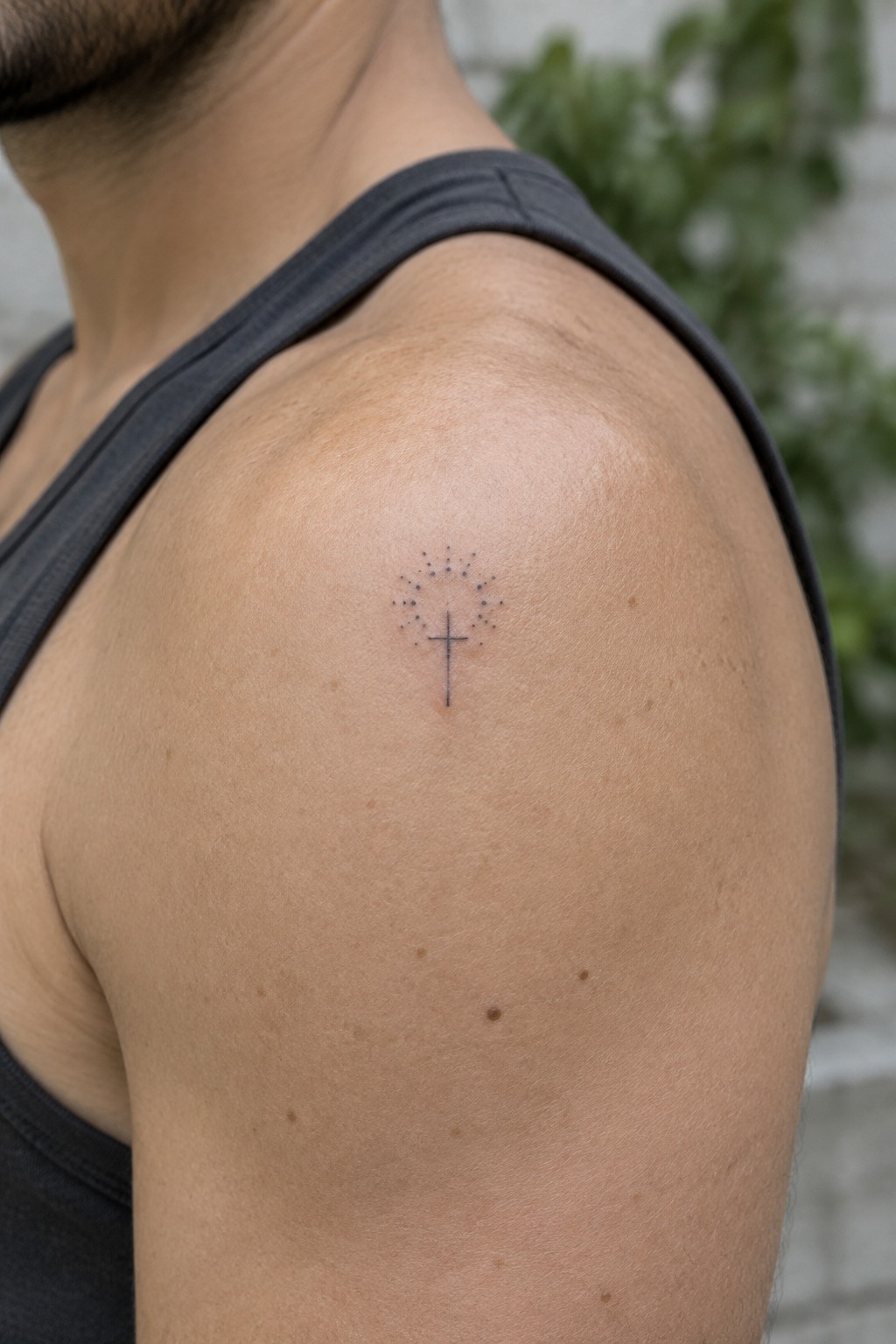 Man's shoulder in gray tank top with small cross and dotted tattoo.