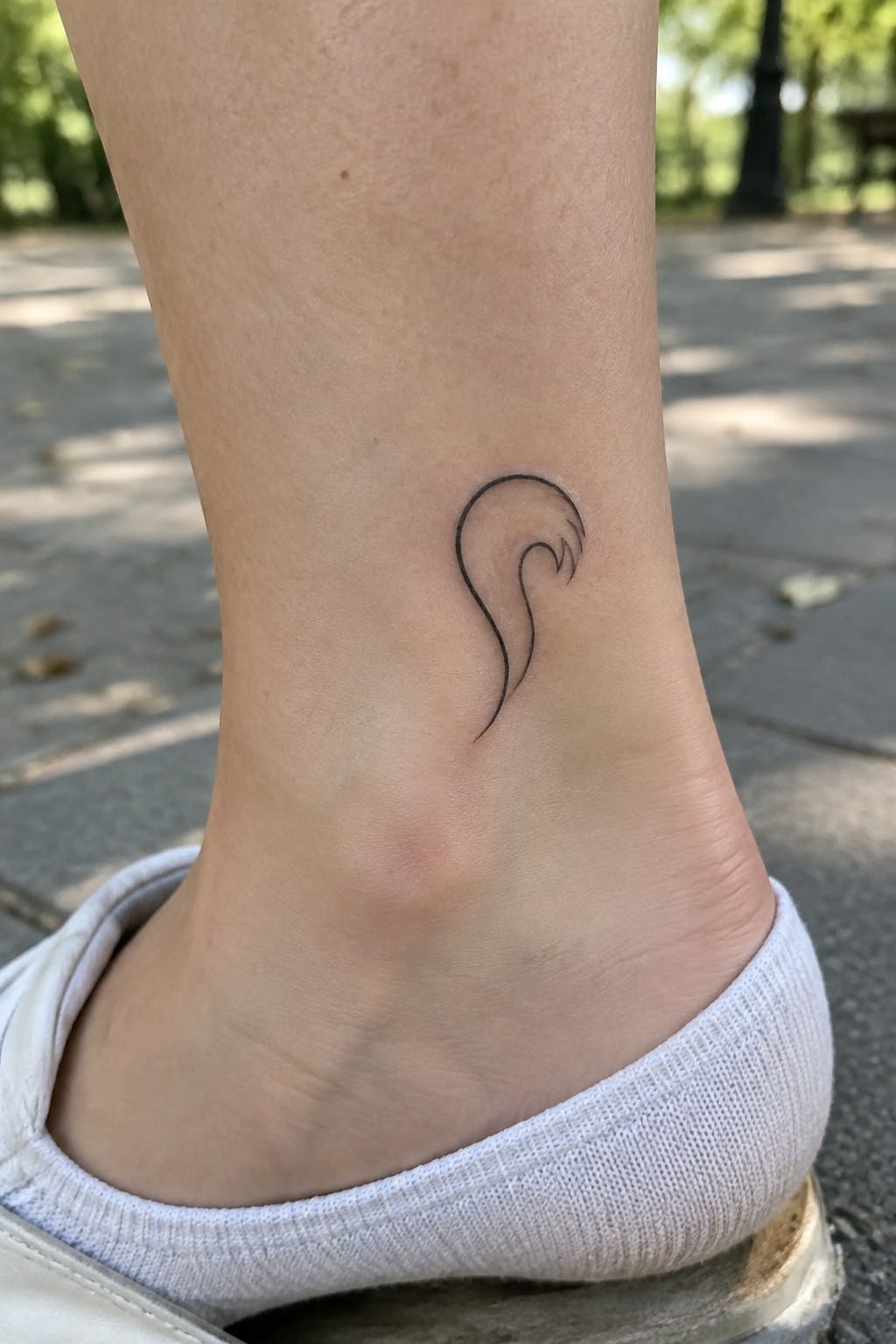 Black swirling line tattoo on ankle above white sock and sneaker on pavement.