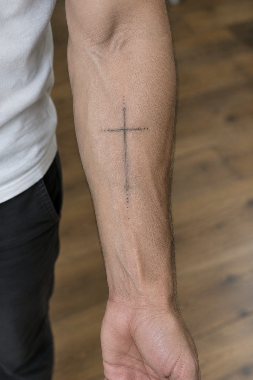 Arm with black dotted cross tattoo, white t-shirt sleeve, wooden floor background.