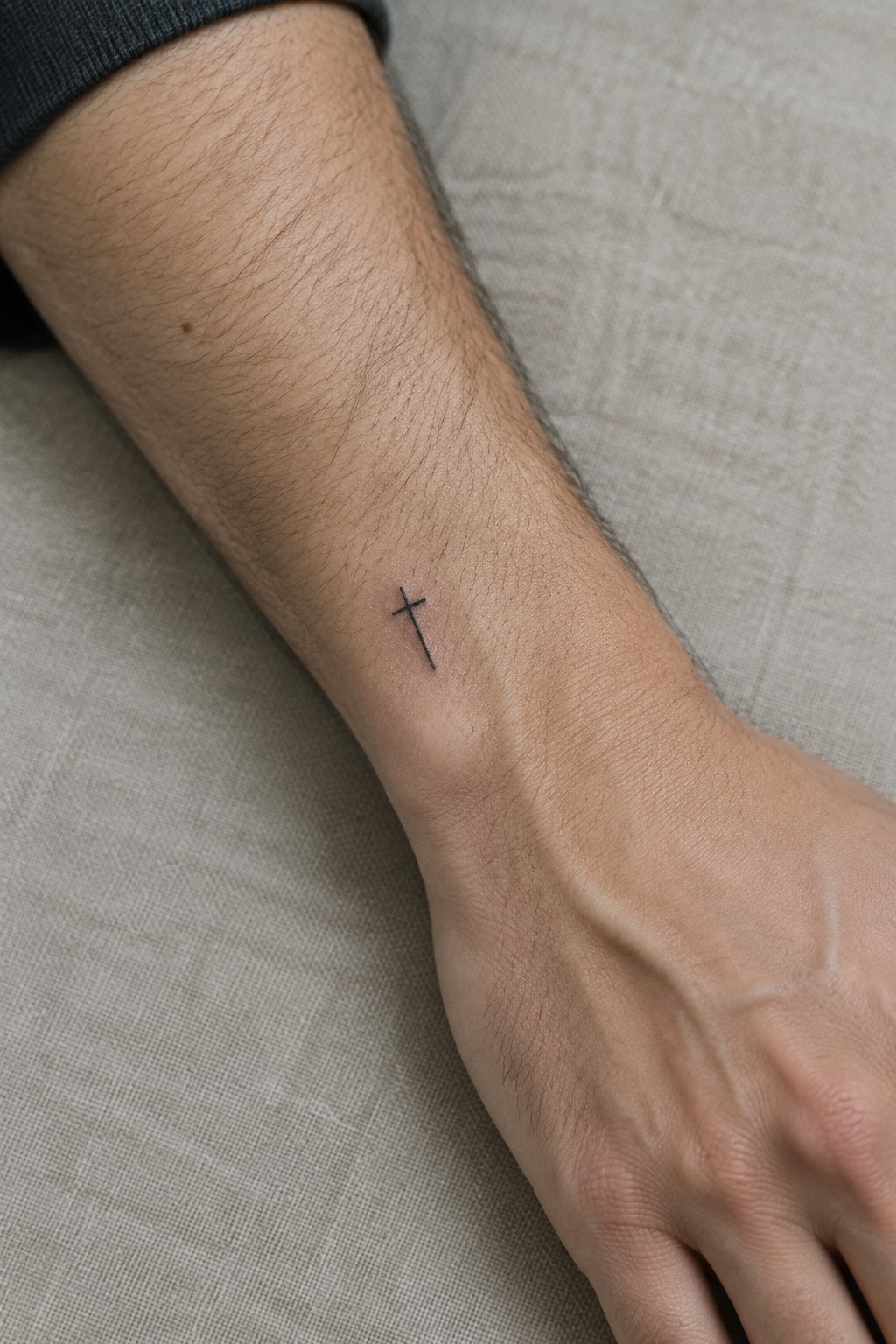 Small black cross tattoo on wrist of arm with dark gray sleeve on beige fabric