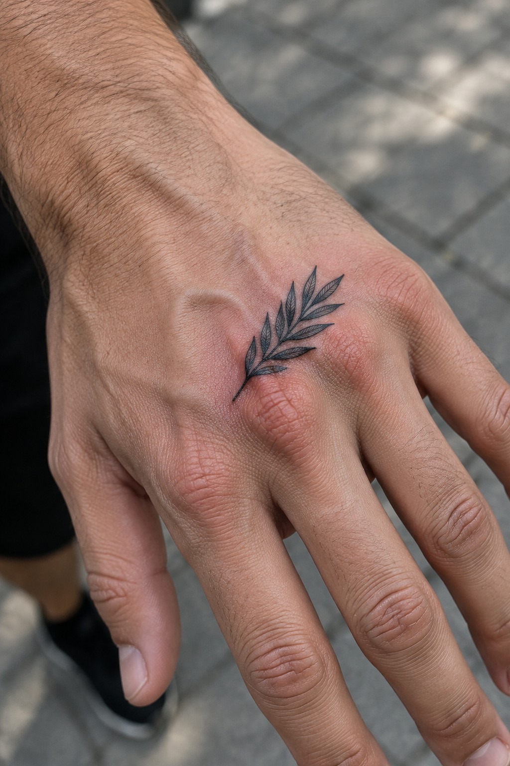Hairy male hand with black leafy branch tattoo on back near fingers.