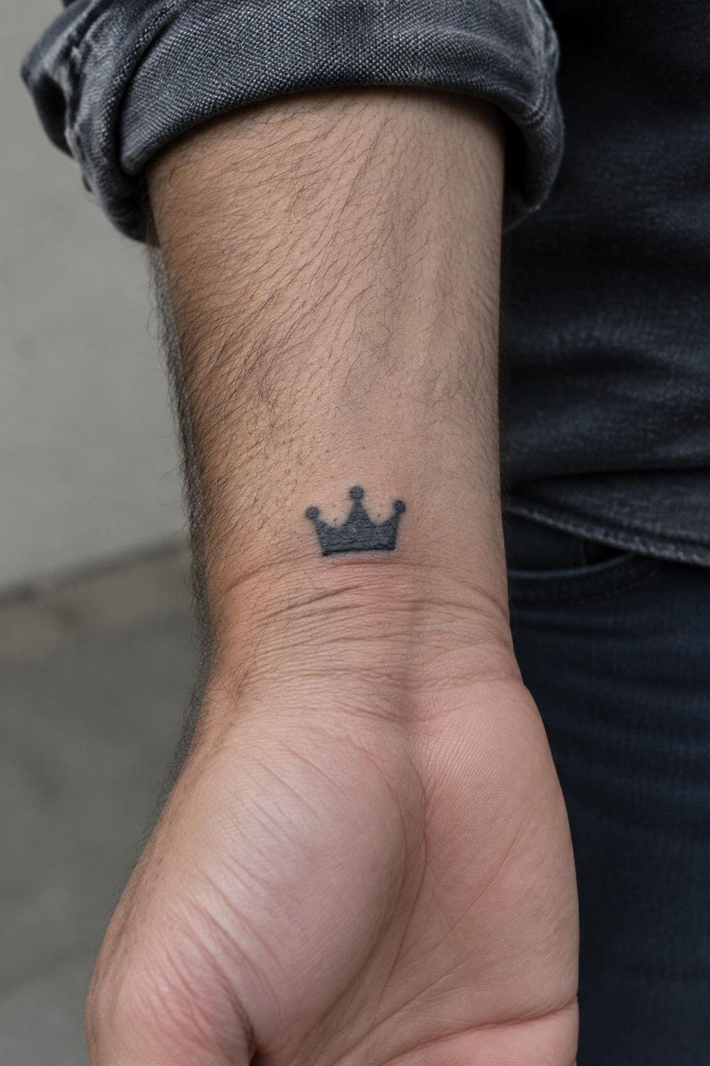 Small black crown tattoo on hairy wrist below rolled gray sleeve