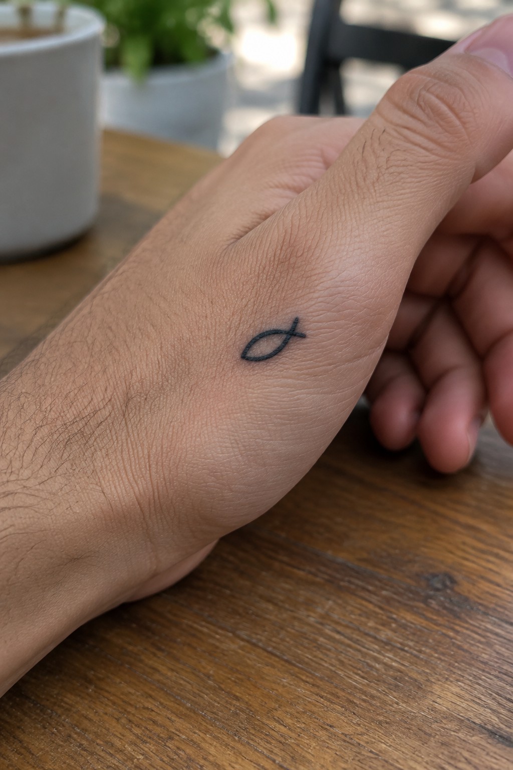 Small black fish tattoo on light-skinned hand resting on wooden table