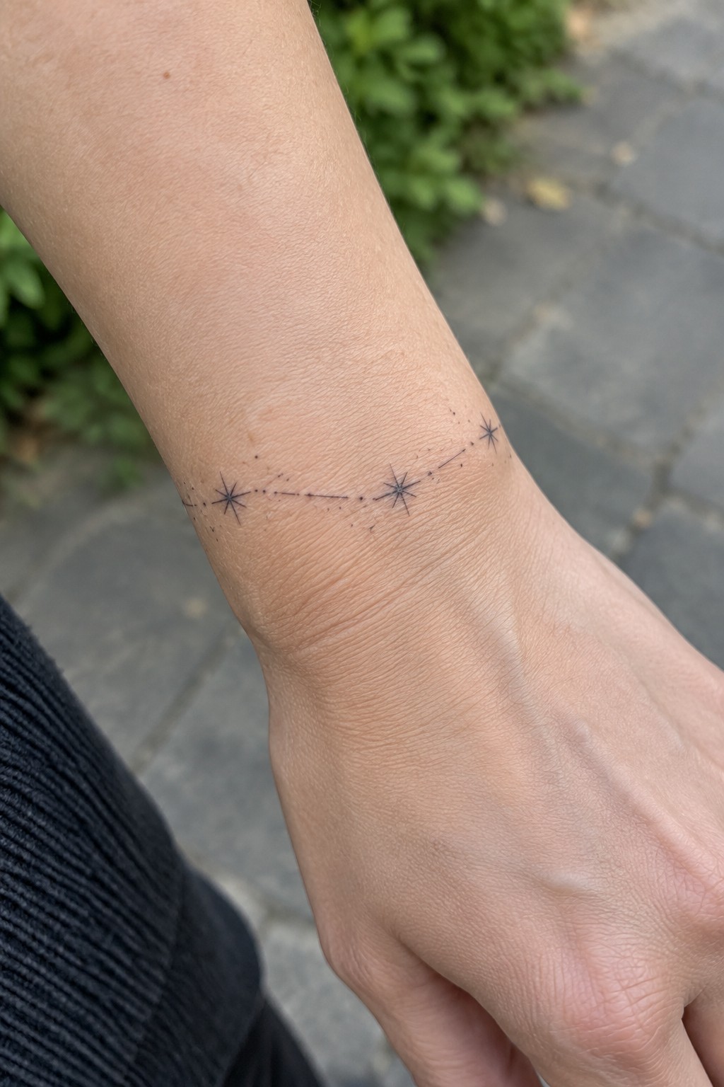 Light-skinned wrist with black constellation tattoo of stars linked by dots, black sleeve visible.