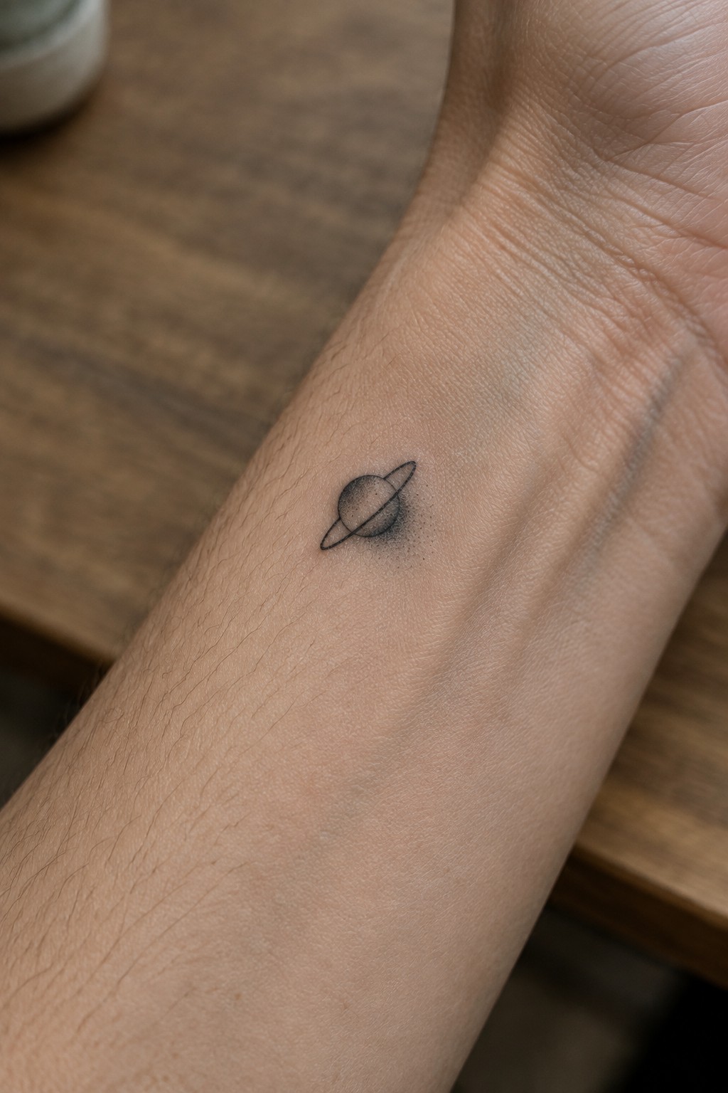 Small fine line tattoo of a planet with rings on the inner wrist in black ink.