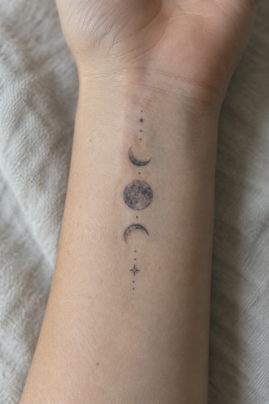 Single needle tattoo of moon phases in a vertical line on the inner wrist with connecting dots and a small star at the bottom