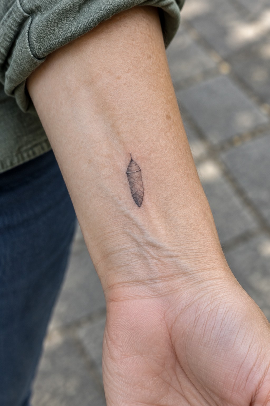 Small chrysalis tattoo on inner wrist below rolled green sleeve and blue jeans