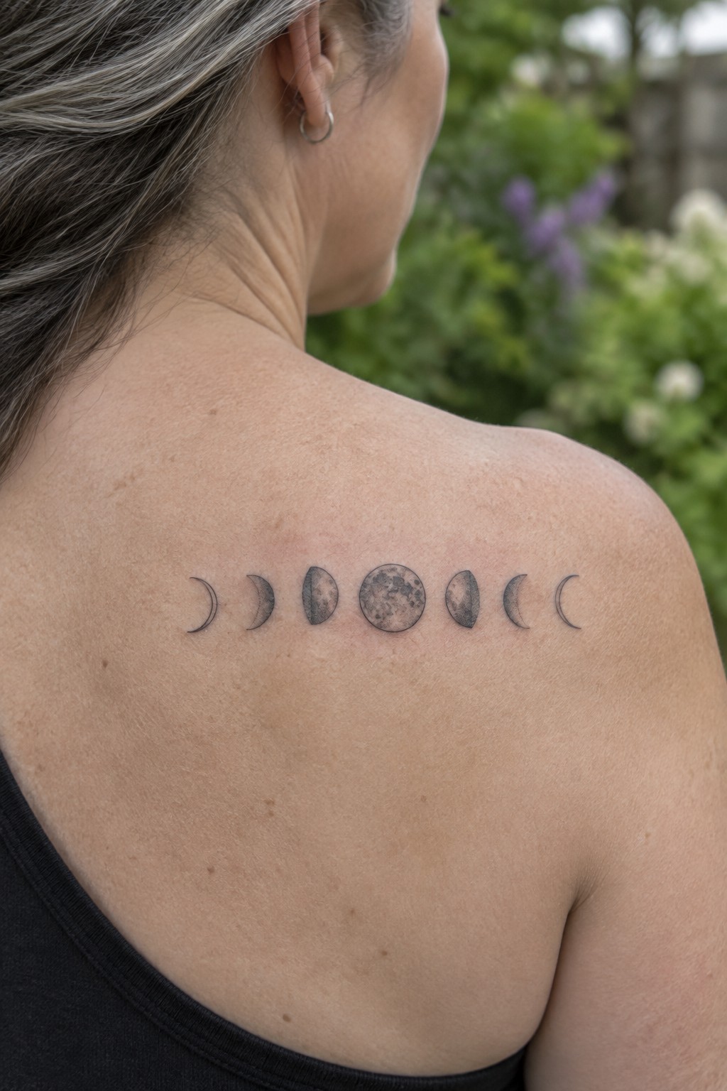 Moon phases tattoo across shoulder above black tank top on woman with graying hair