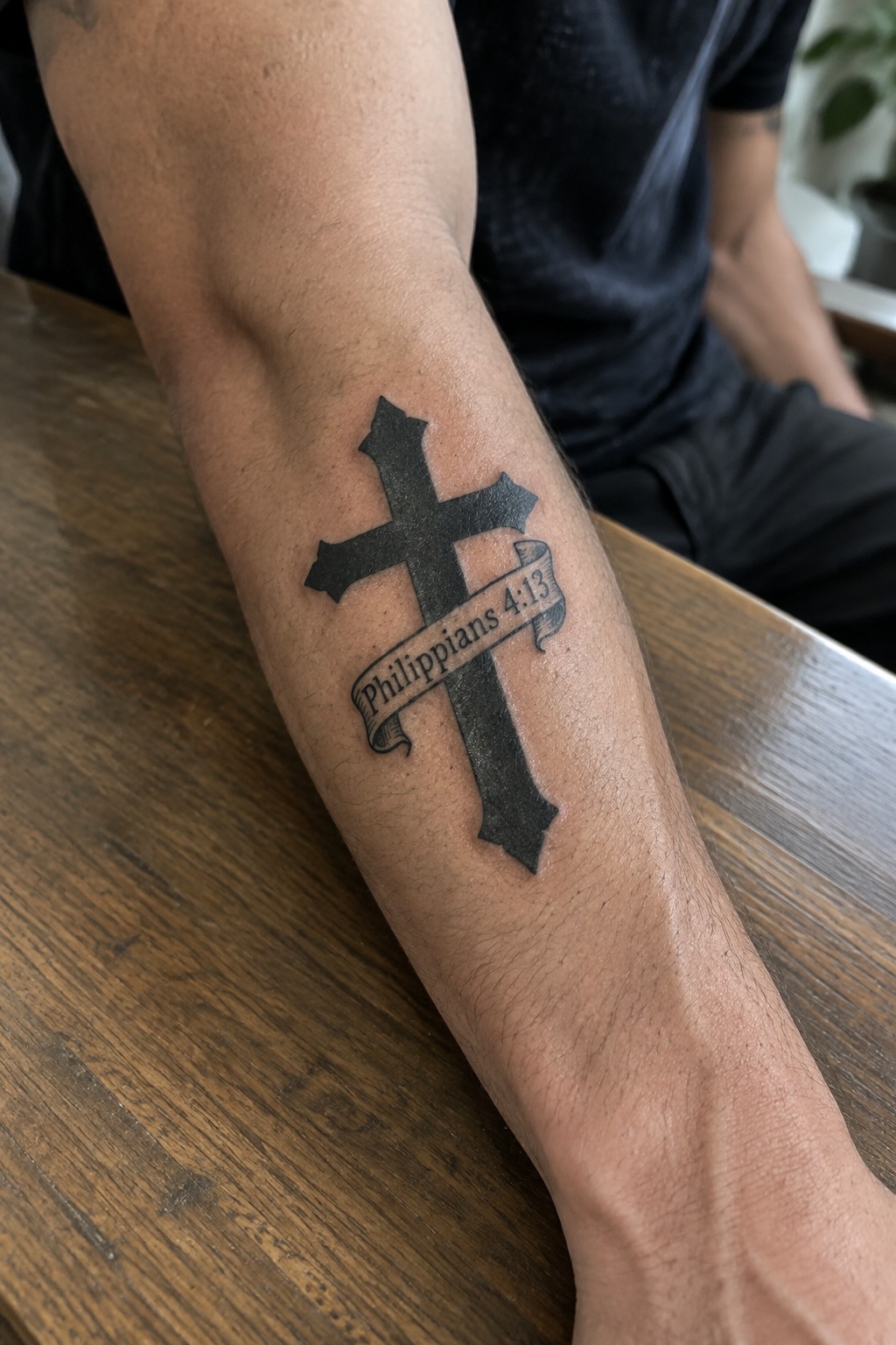 Black cross tattoo with a Philippians 4:13 banner on a man's forearm