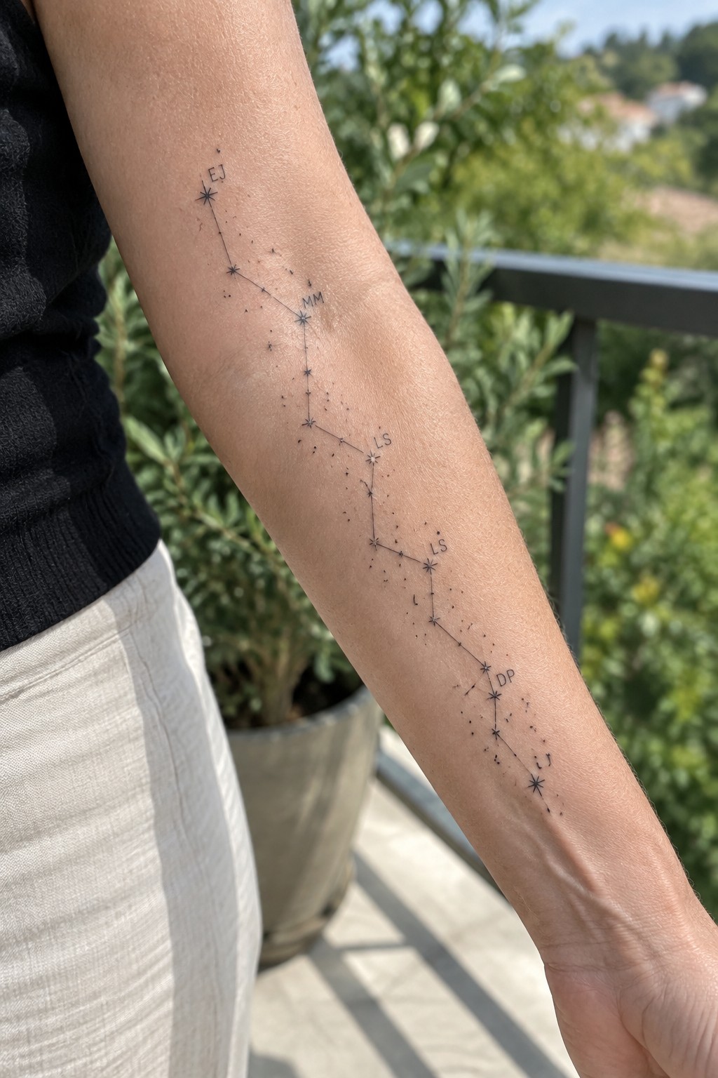 Arm with constellation tattoo of connected stars and initials, black top, white pants outdoors.