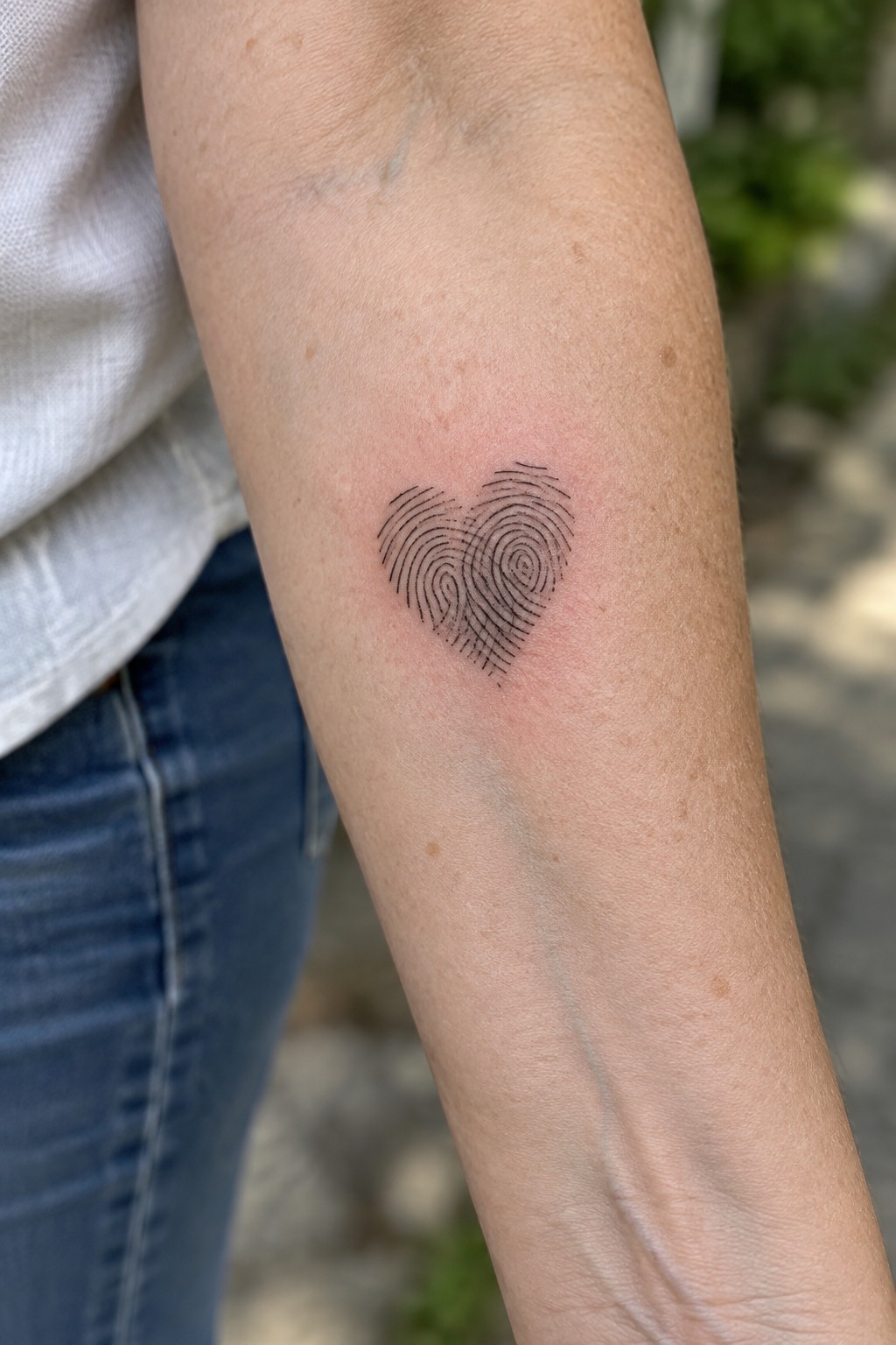 Fresh fingerprint heart tattoo on reddened forearm beside white shirt and blue jeans.