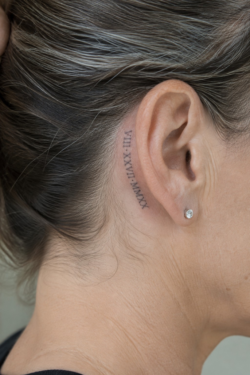Dark hair with gray strands and Roman numeral tattoo behind ear with diamond stud earring