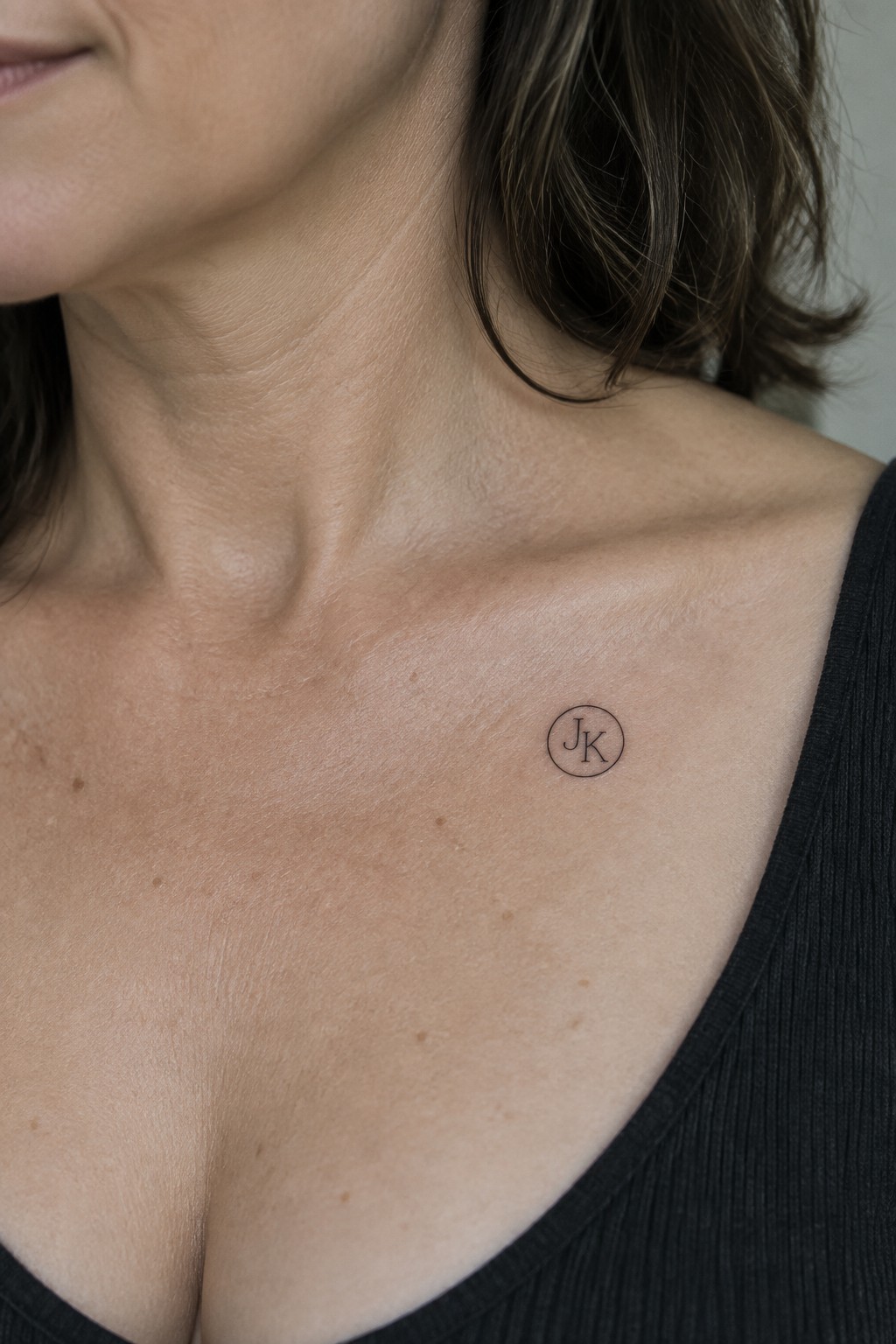 Circular JK tattoo on woman's freckled collarbone above black ribbed top