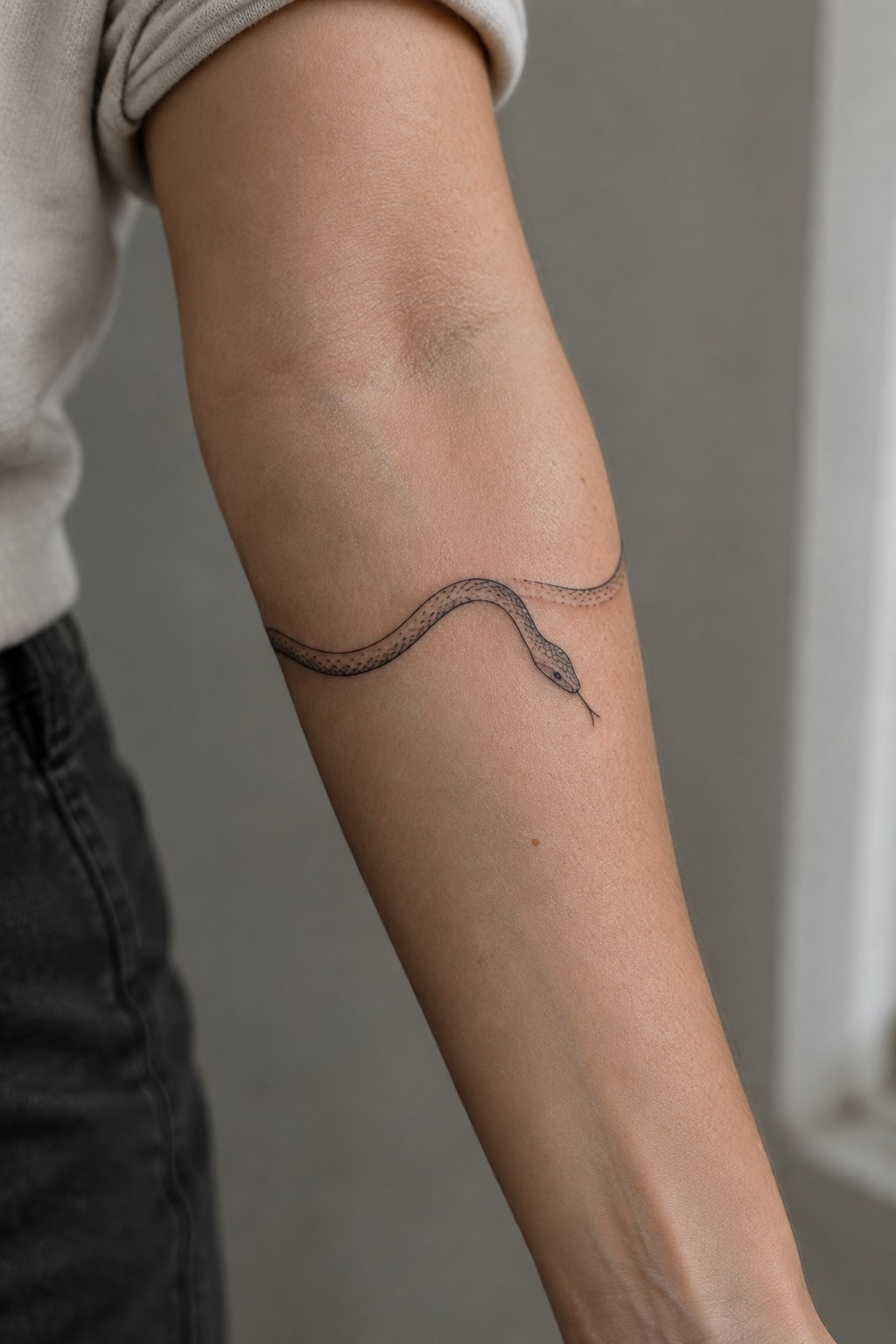 Arm with black snake tattoo, beige shirt sleeve, dark pants, gray background.