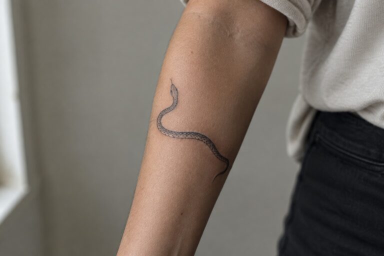 Arm with black snake tattoo, beige shirt sleeve, dark pants, gray background.