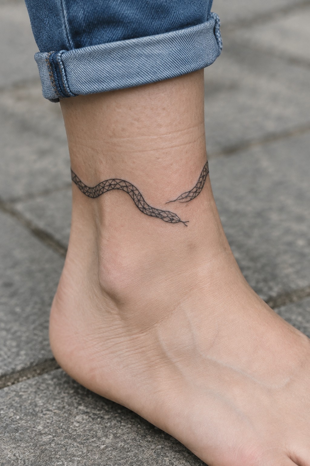 Bare foot with geometric black snake tattoo on ankle, blue jeans rolled up.