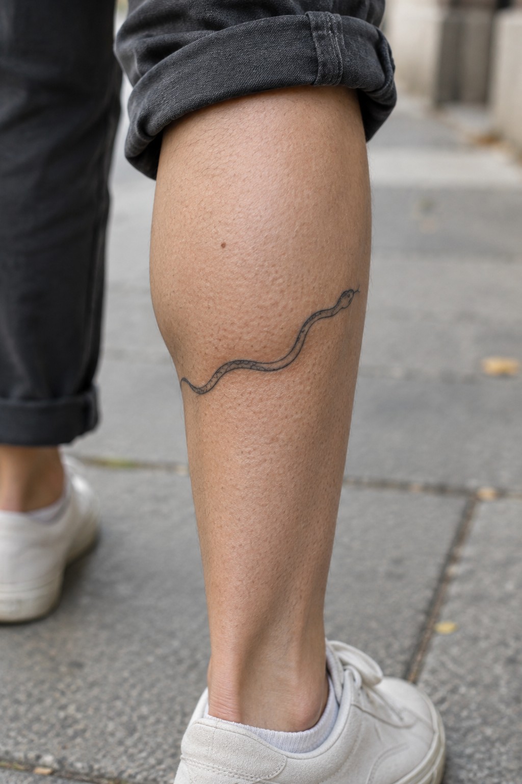Snake tattoo on calf below rolled gray pants and white sneakers on pavement