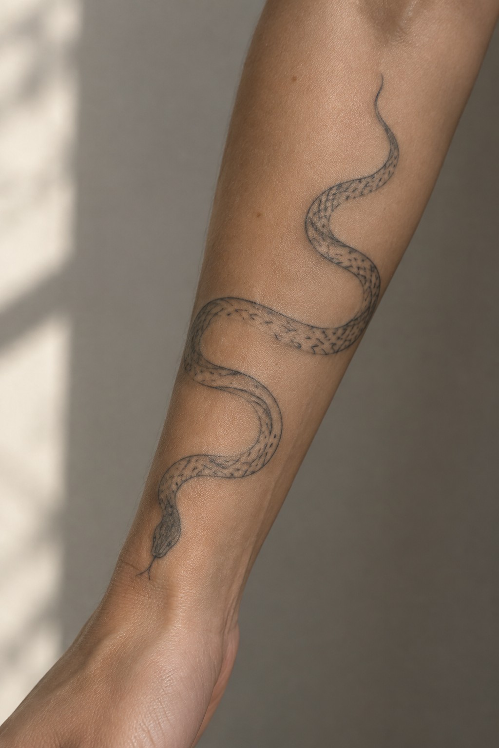 Fine line snake tattoo curving along a woman's forearm with the head near the wrist and scales lightly shaded.