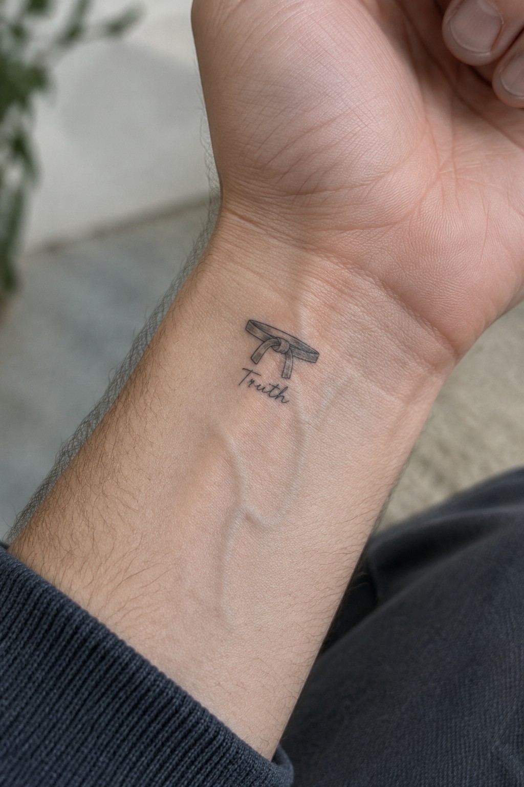 Inner wrist tattoo of tied martial arts belt above cursive word Truth