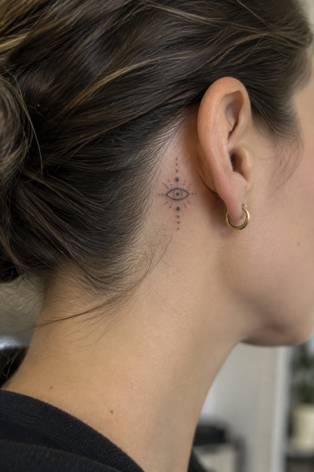 Woman's neck with eye tattoo behind ear and small gold hoop earring, hair in bun.