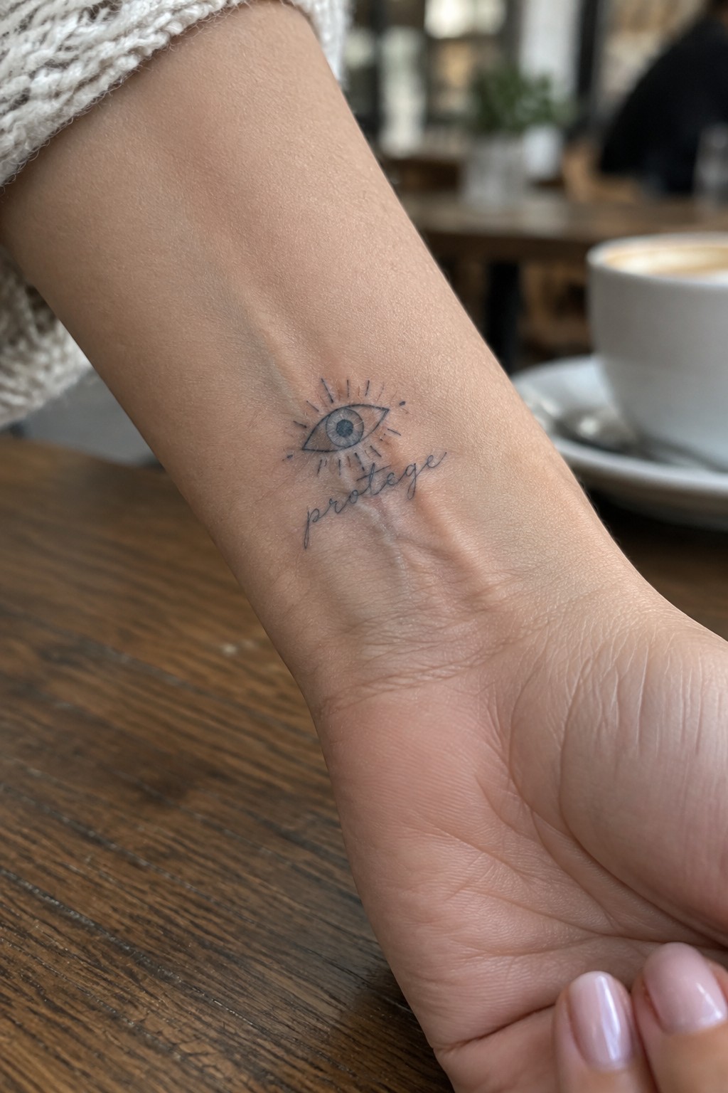 Small fine line evil eye tattoo with rays and the word protege placed on the inner wrist.
