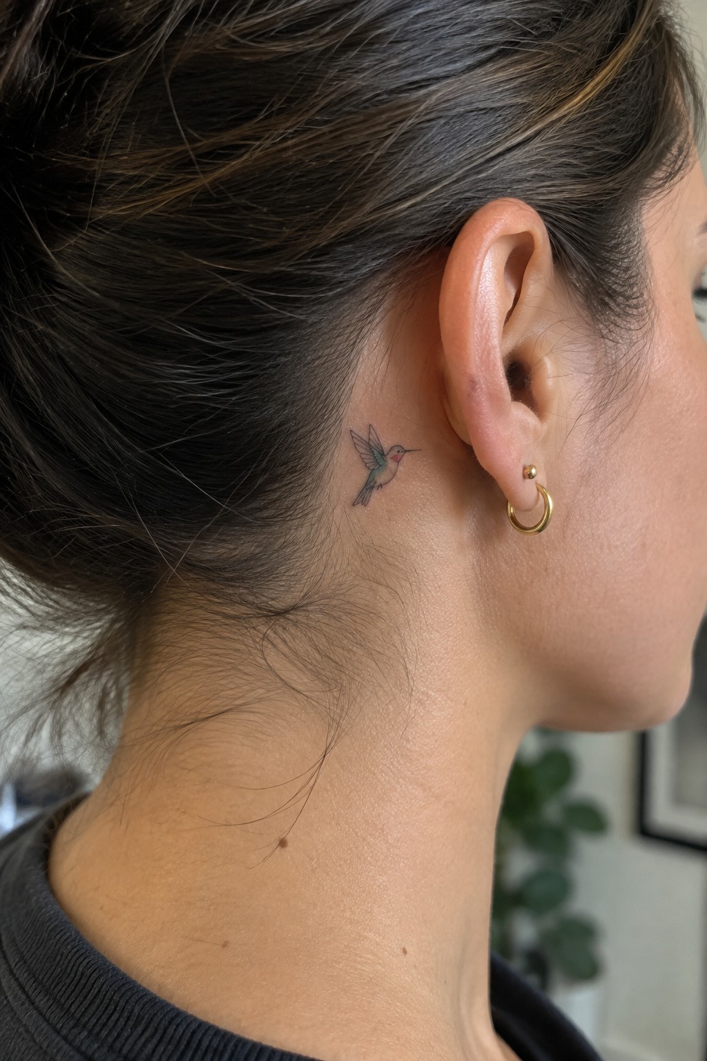Woman with dark bun, small hummingbird tattoo behind ear, gold hoop earring