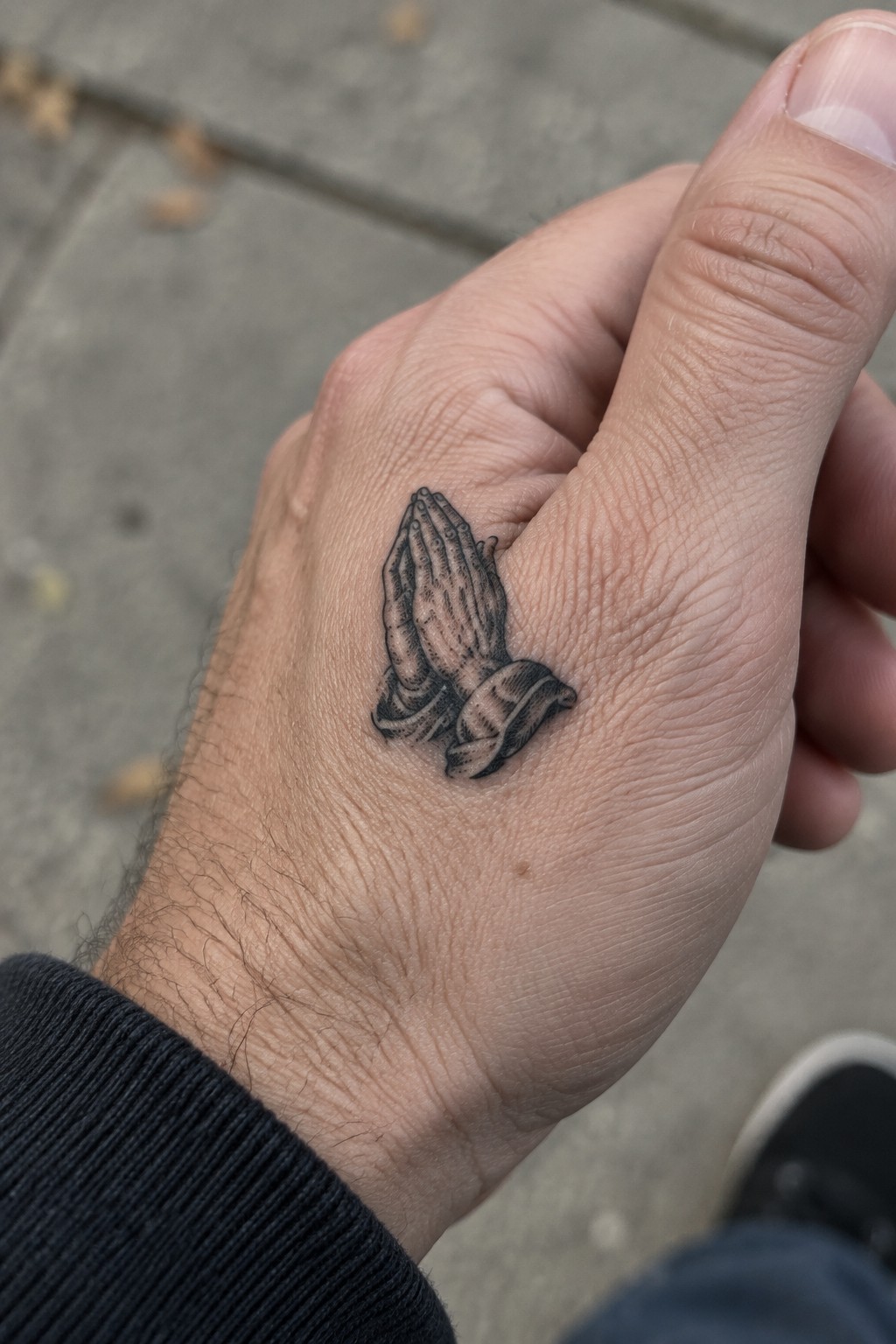 Small engraving style praying hands tattoo on the back of a man's hand with a ribbon detail below