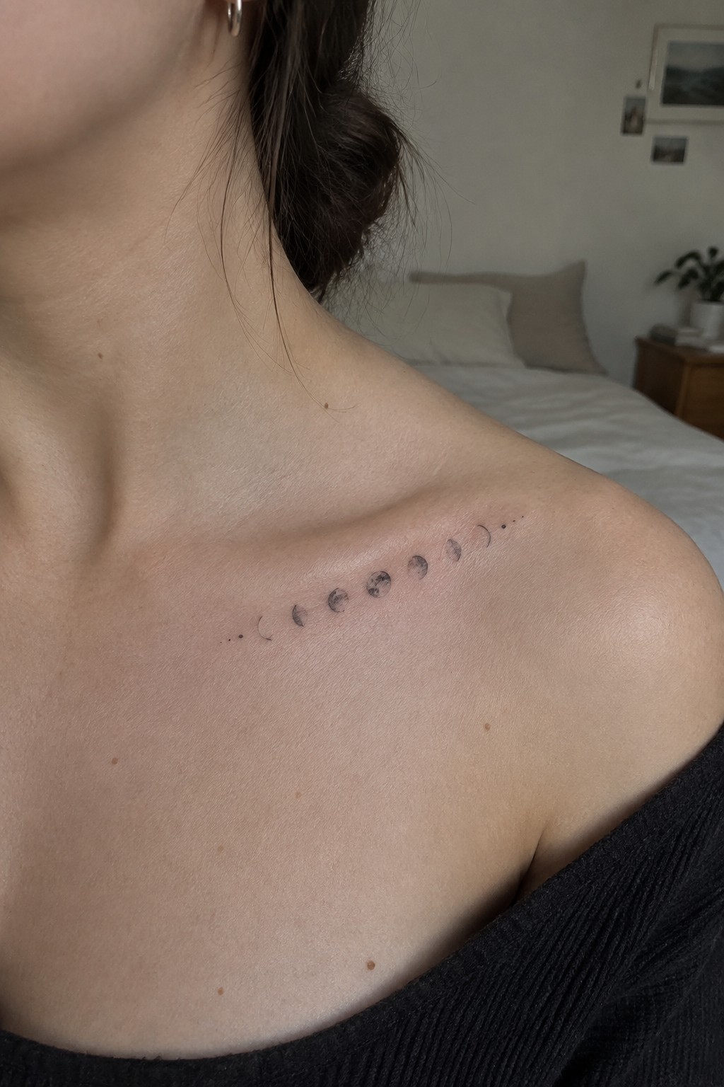 Moon phase tattoo on shoulder above black off-shoulder top in bedroom
