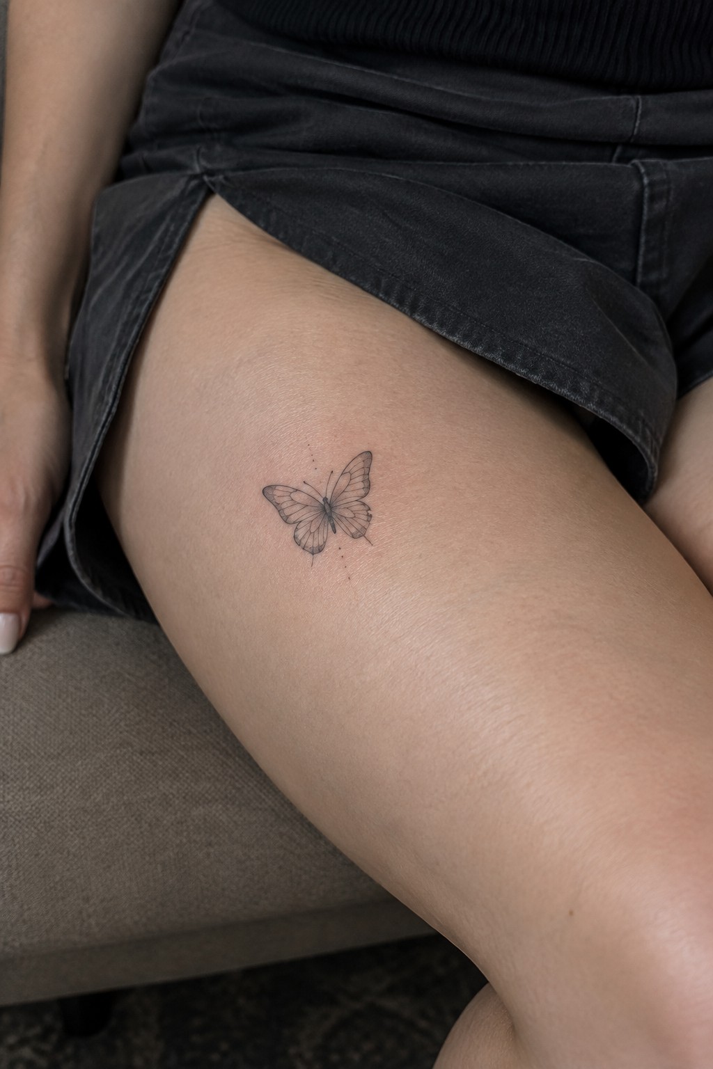 Delicate black butterfly tattoo on thigh under dark gray denim shorts.