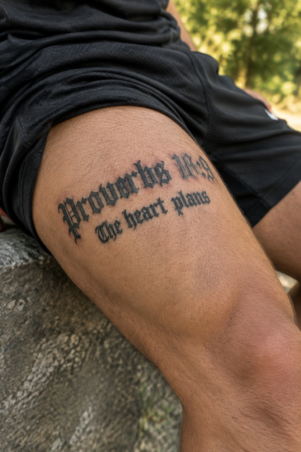 Close up of a man's thigh showing a Proverbs 16:9 tattoo in bold gothic lettering with the words The heart plans below it